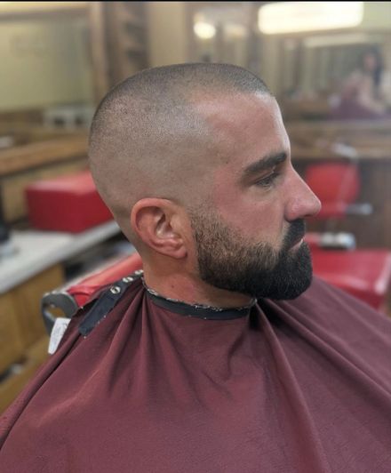 Man with a faded buzz cut and beard, wearing a barber's cape, seated in a shop.