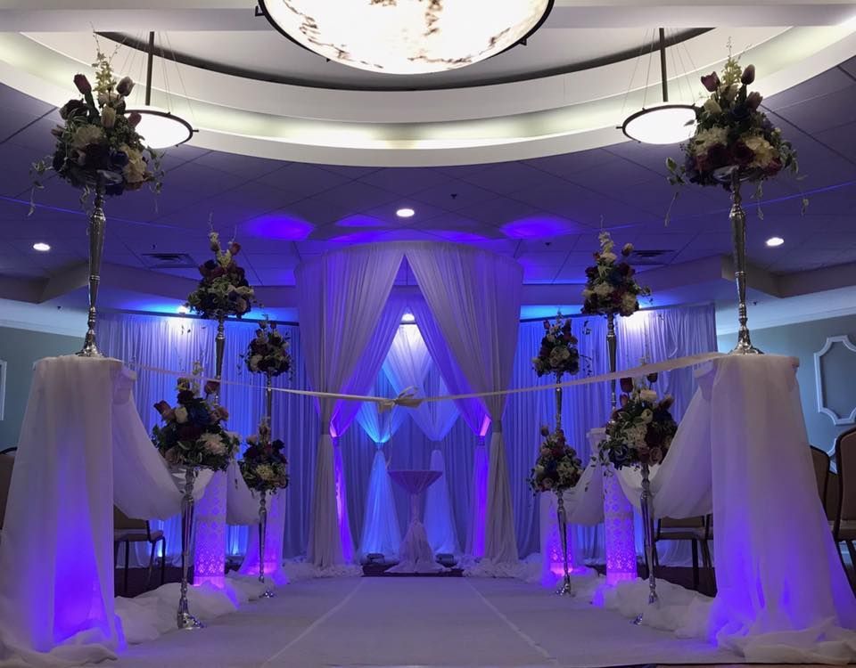 Elegant wedding venue with purple uplighting, draped white curtains, and floral arrangements on pedestals.
