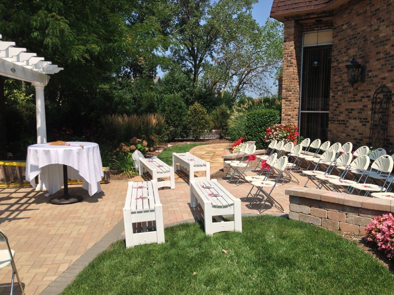 Patio with white benches, round table, and rows of white chairs beside a brick house and garden