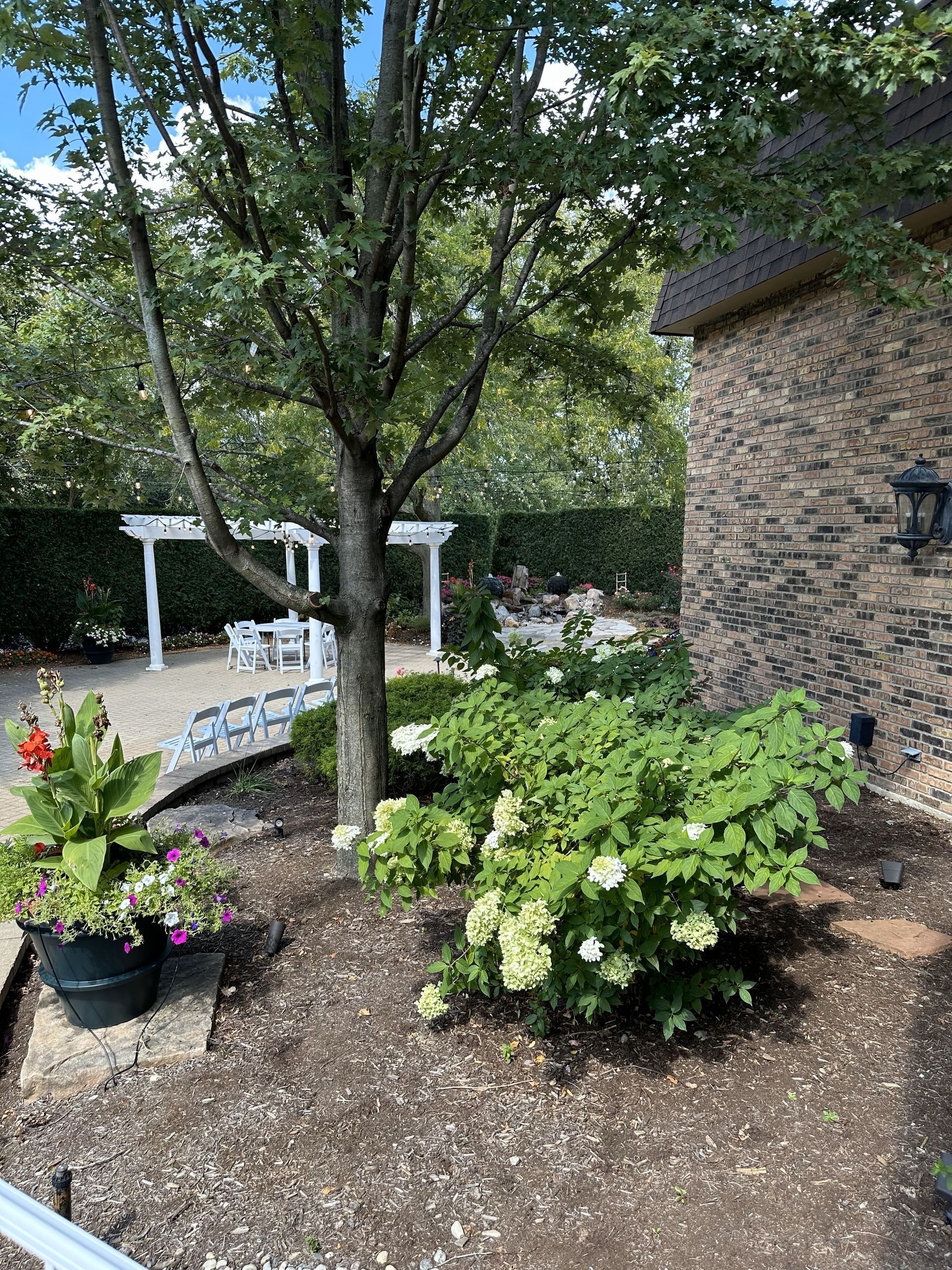 Garden patio with a large tree, potted flowers, and white outdoor seating beside a stone house.