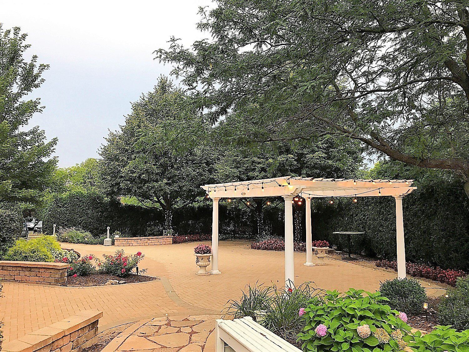 Garden path with a white pergola, flowering shrubs, and trees under a cloudy sky