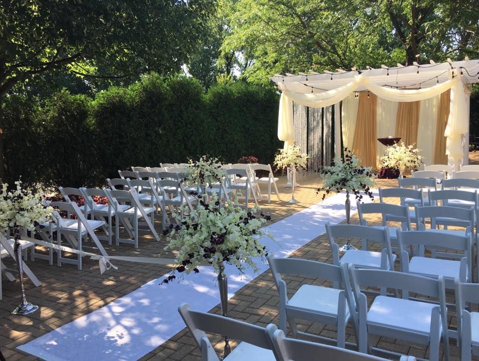 Outdoor wedding ceremony setup with white chairs, aisle runner, and draped canopy in a garden