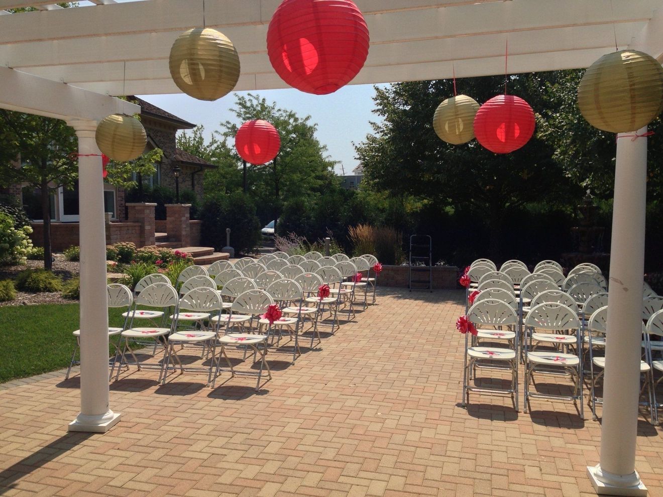 Outdoor wedding ceremony setup with rows of white chairs under red and gold lanterns on a brick patio
