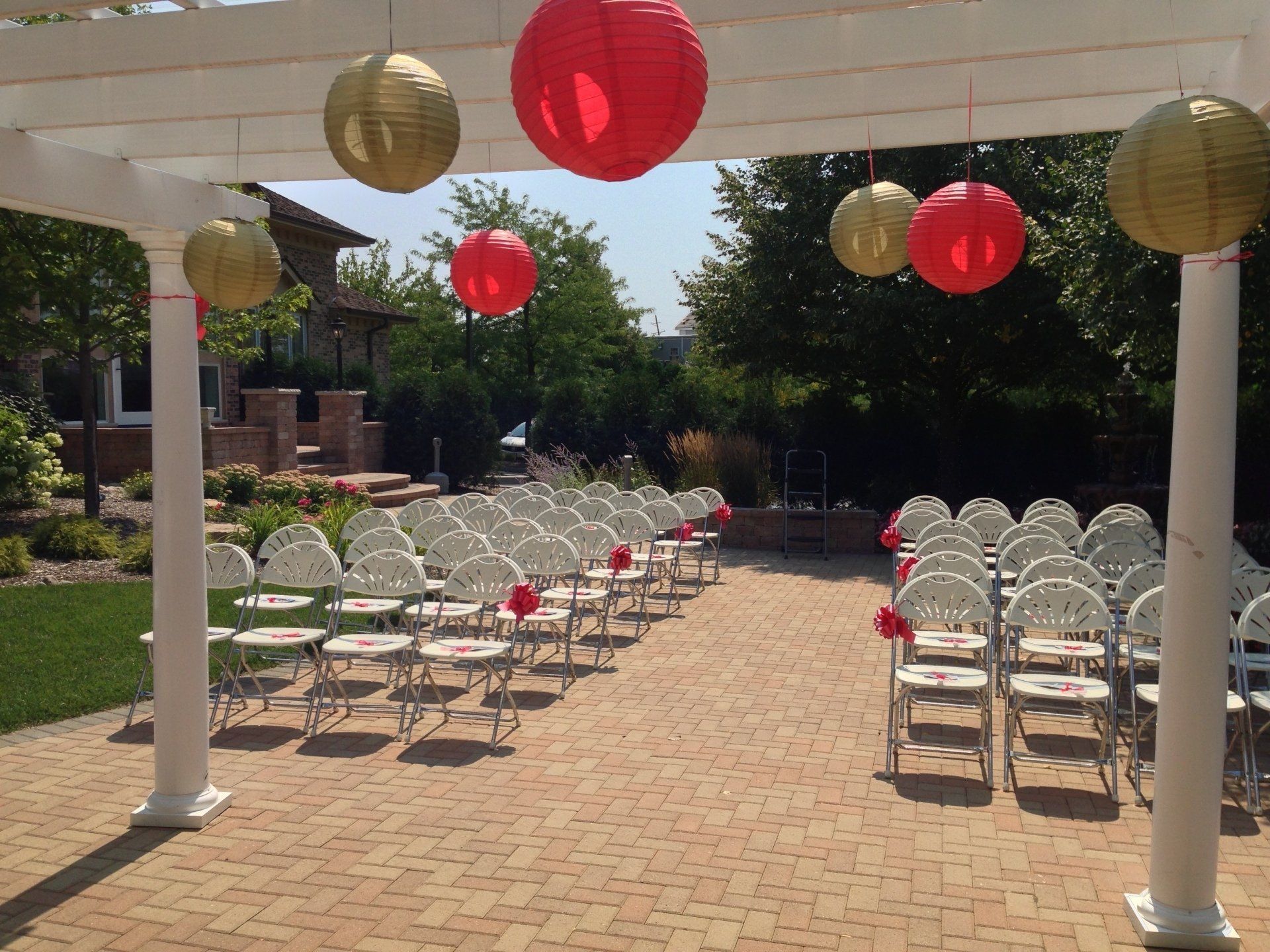 Outdoor wedding ceremony setup with rows of white chairs under red and gold lanterns on a brick patio