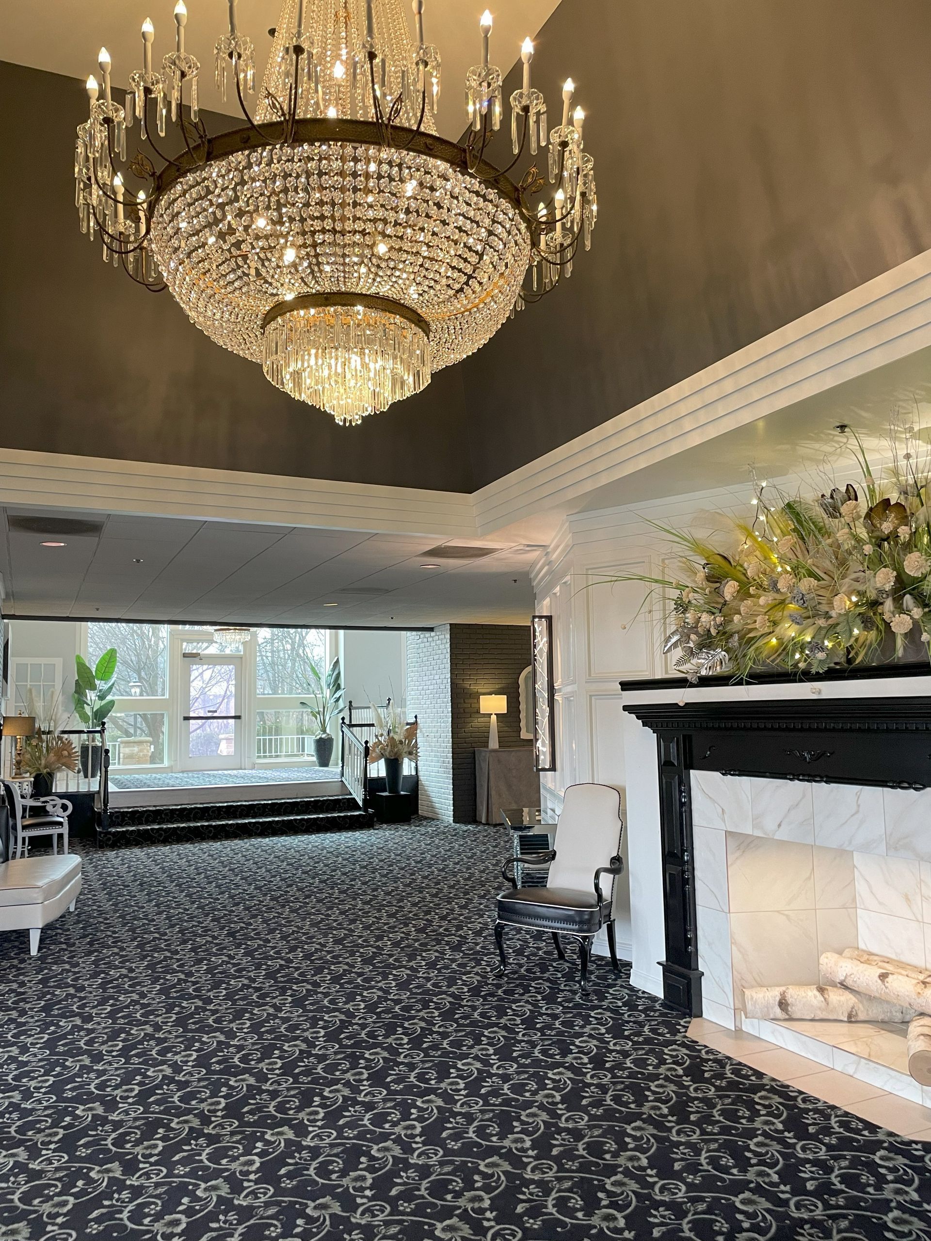 Elegant lobby with crystal chandelier, black floral carpet, white fireplace, and large windows