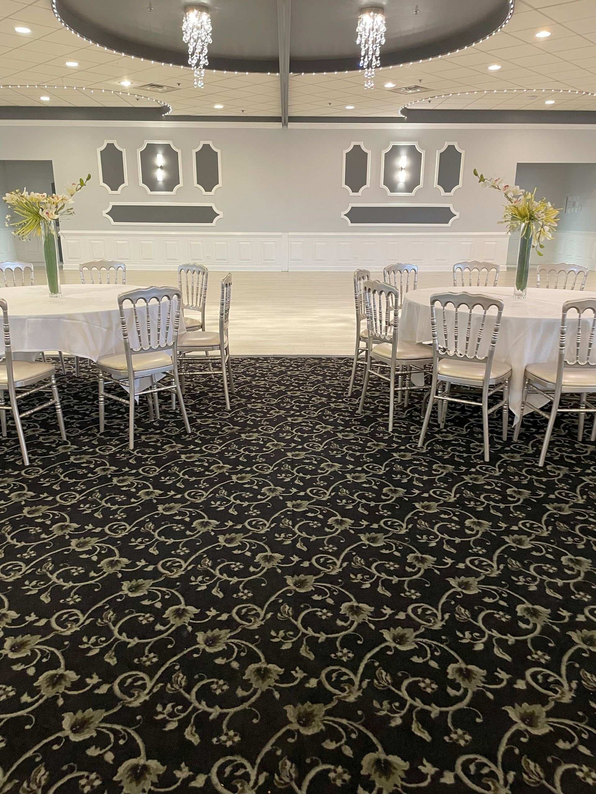 Elegant banquet hall with round tables, white chairs, chandeliers, and a patterned black carpet.