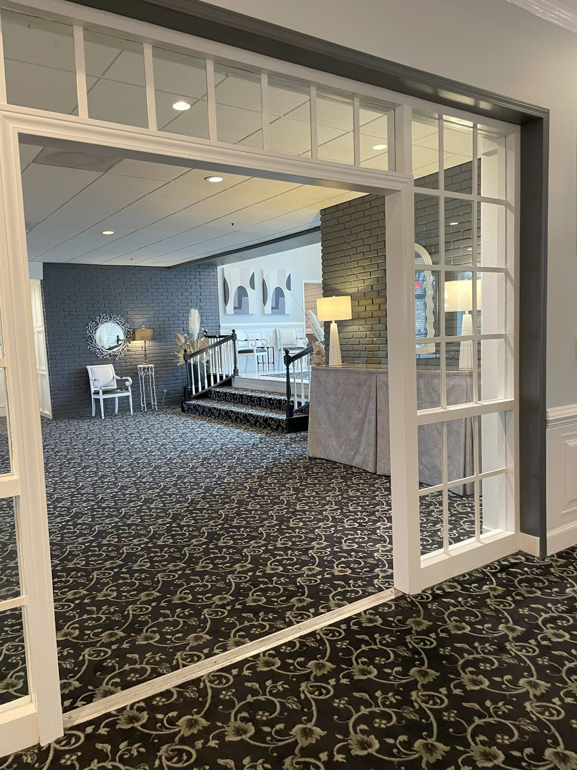 Open doorway to a carpeted lounge with tables, chairs, and a lamp beyond a patterned hall rug.