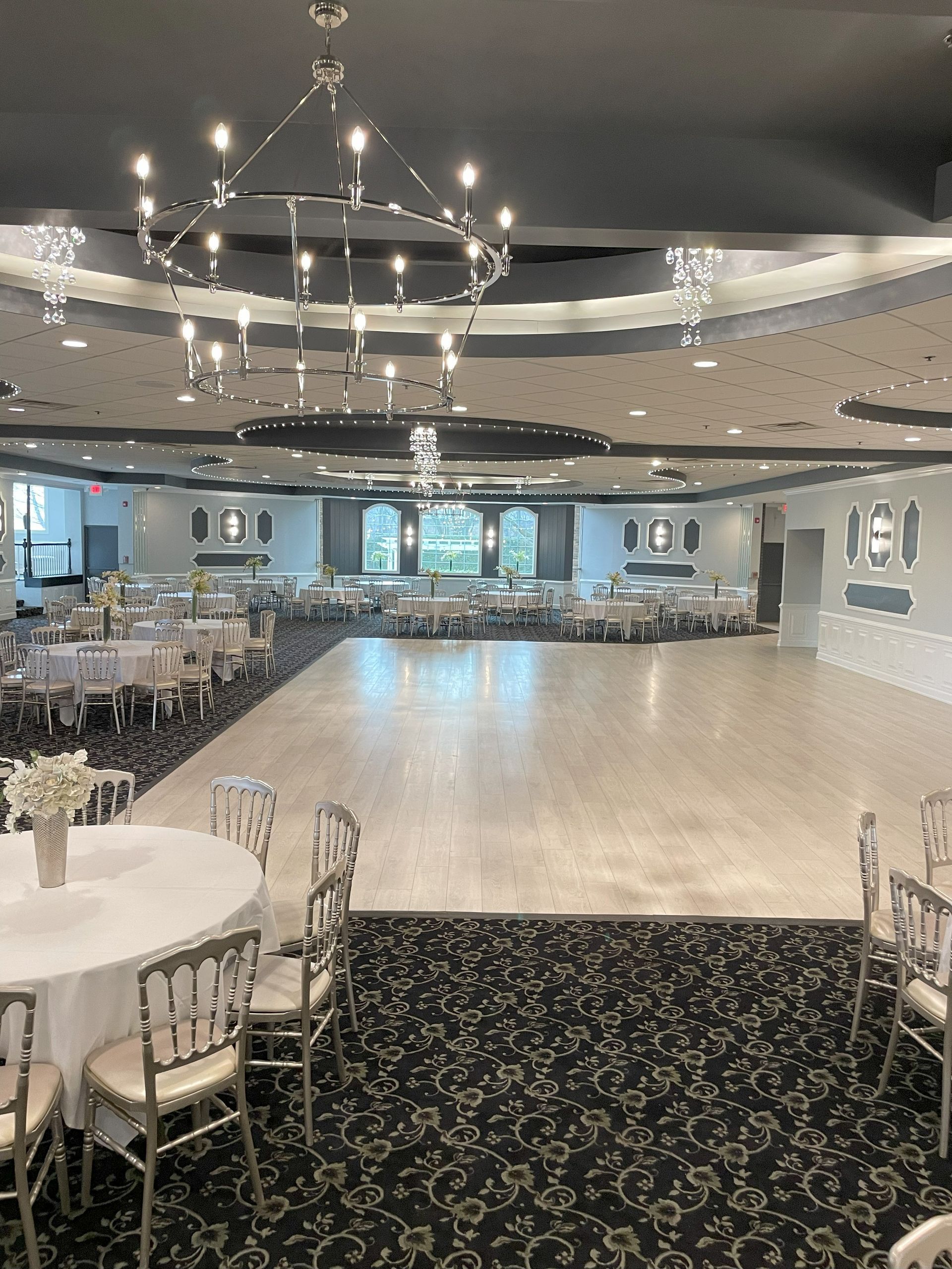 Empty elegant banquet hall with round tables, white chairs, chandeliers, and a light dance floor