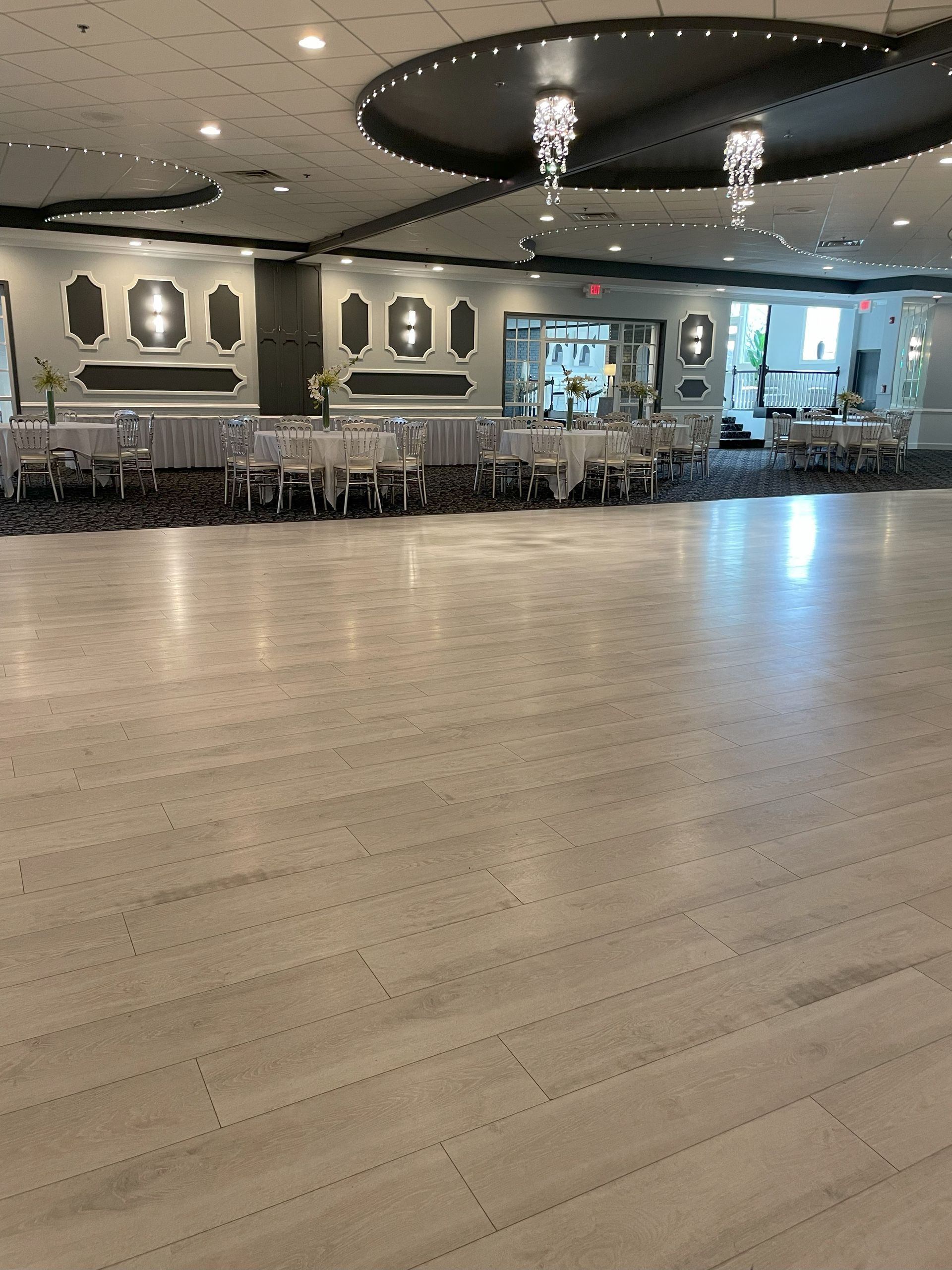 Empty elegant banquet hall with white floors, mirrored walls, chandeliers, and rows of chairs along the back wall