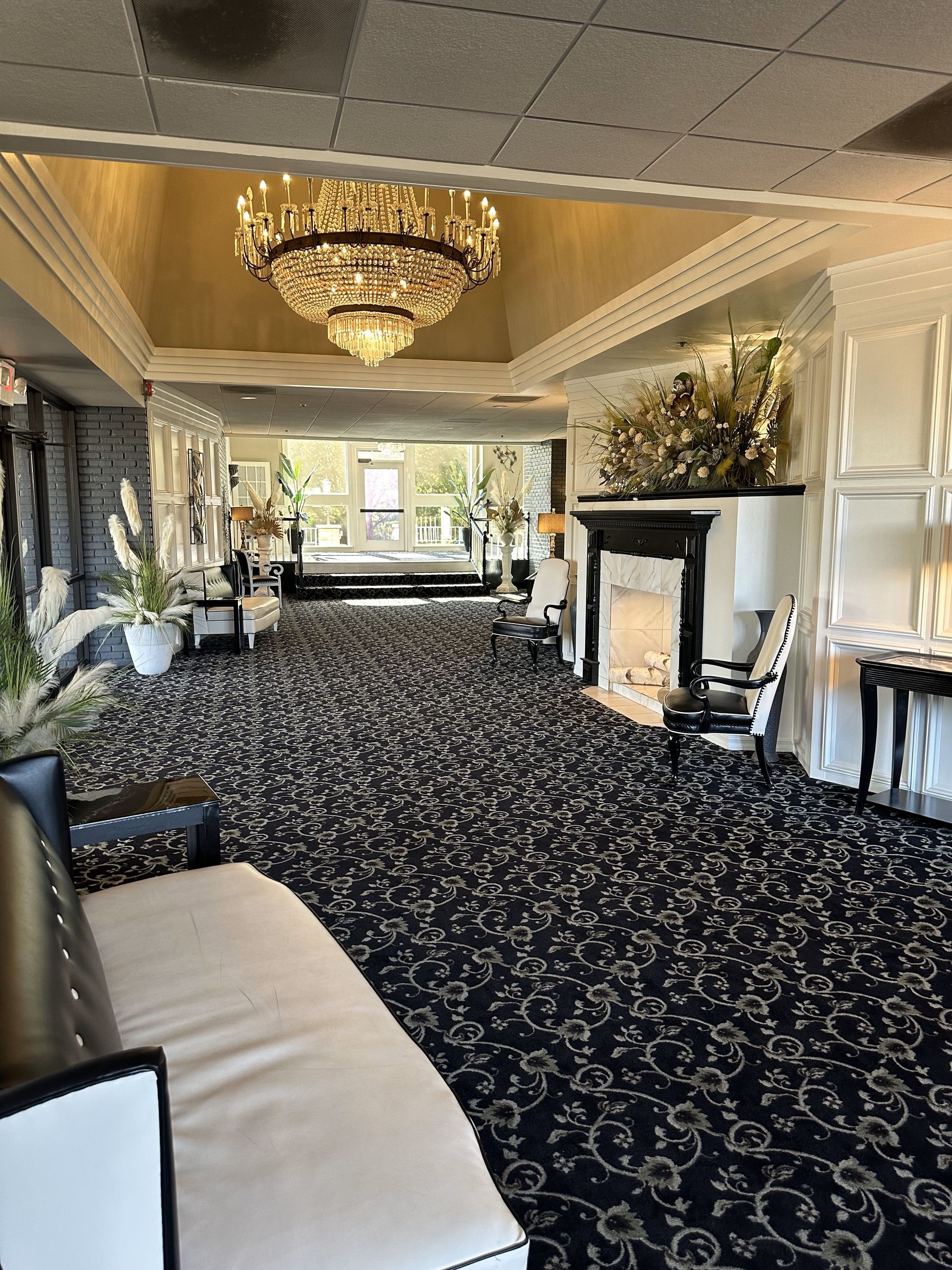 Elegant lounge with chandelier, patterned carpet, fireplace, and white chairs in a bright room