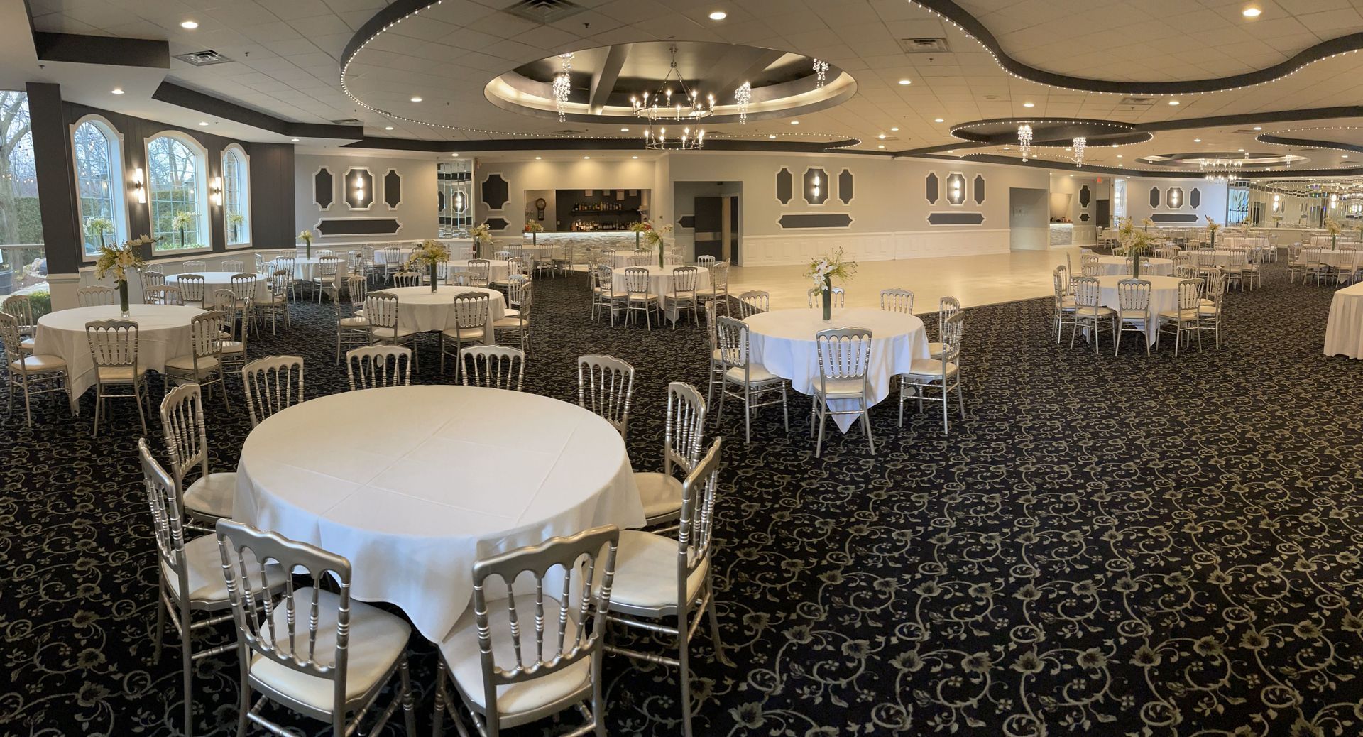 Elegant banquet hall with round tables, white tablecloths, silver chairs, and patterned carpet under chandeliers