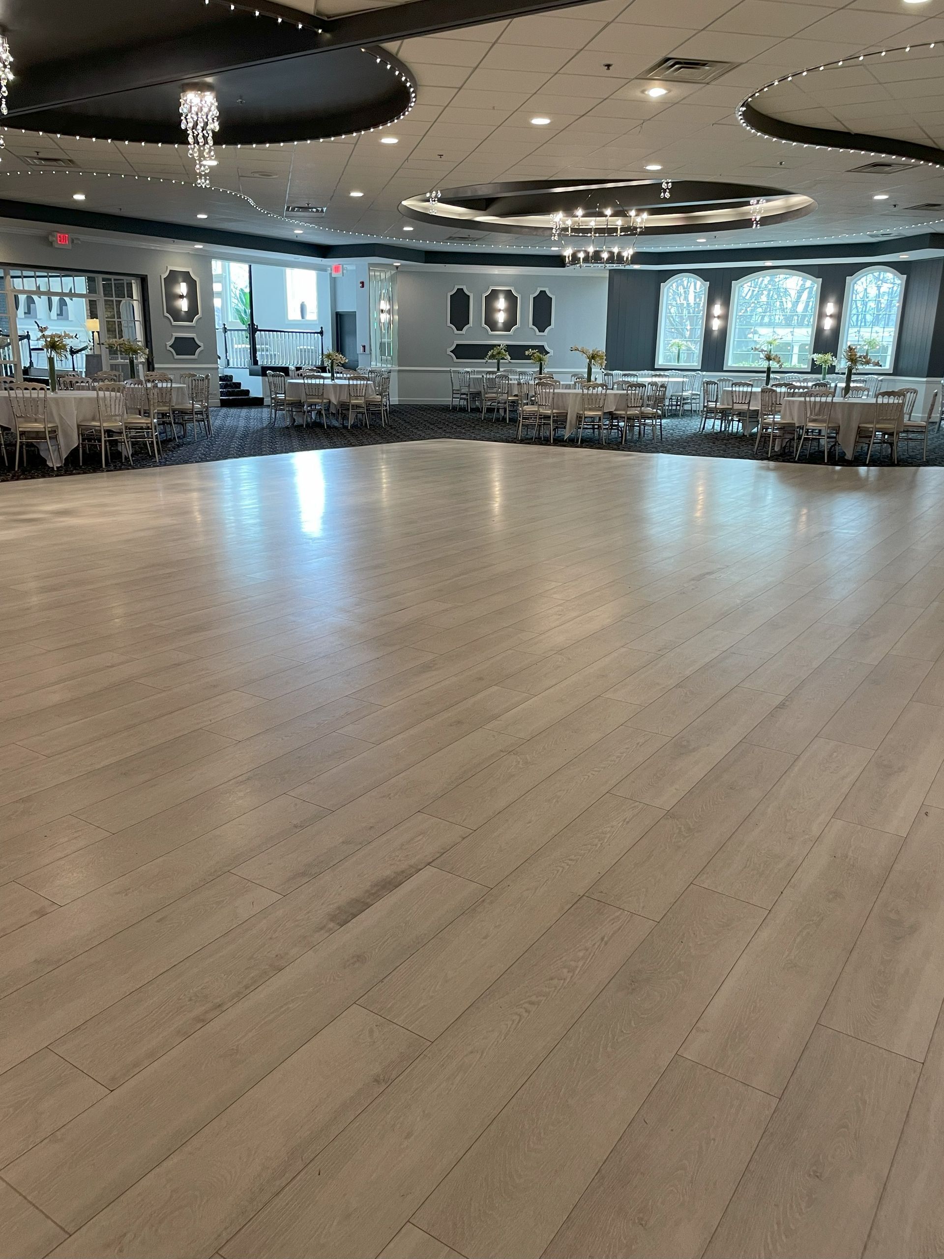 Empty ballroom with polished wooden floor, round ceiling lights, and rows of tables and chairs along the walls