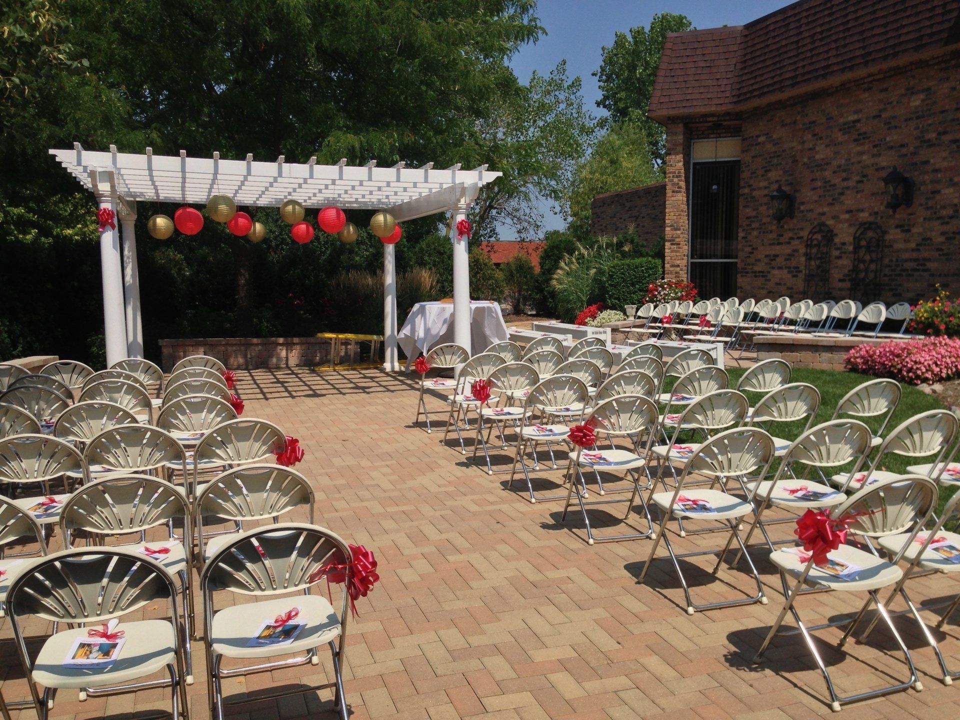 Outdoor event setup with rows of white chairs, a pergola, and red lanterns on a brick patio