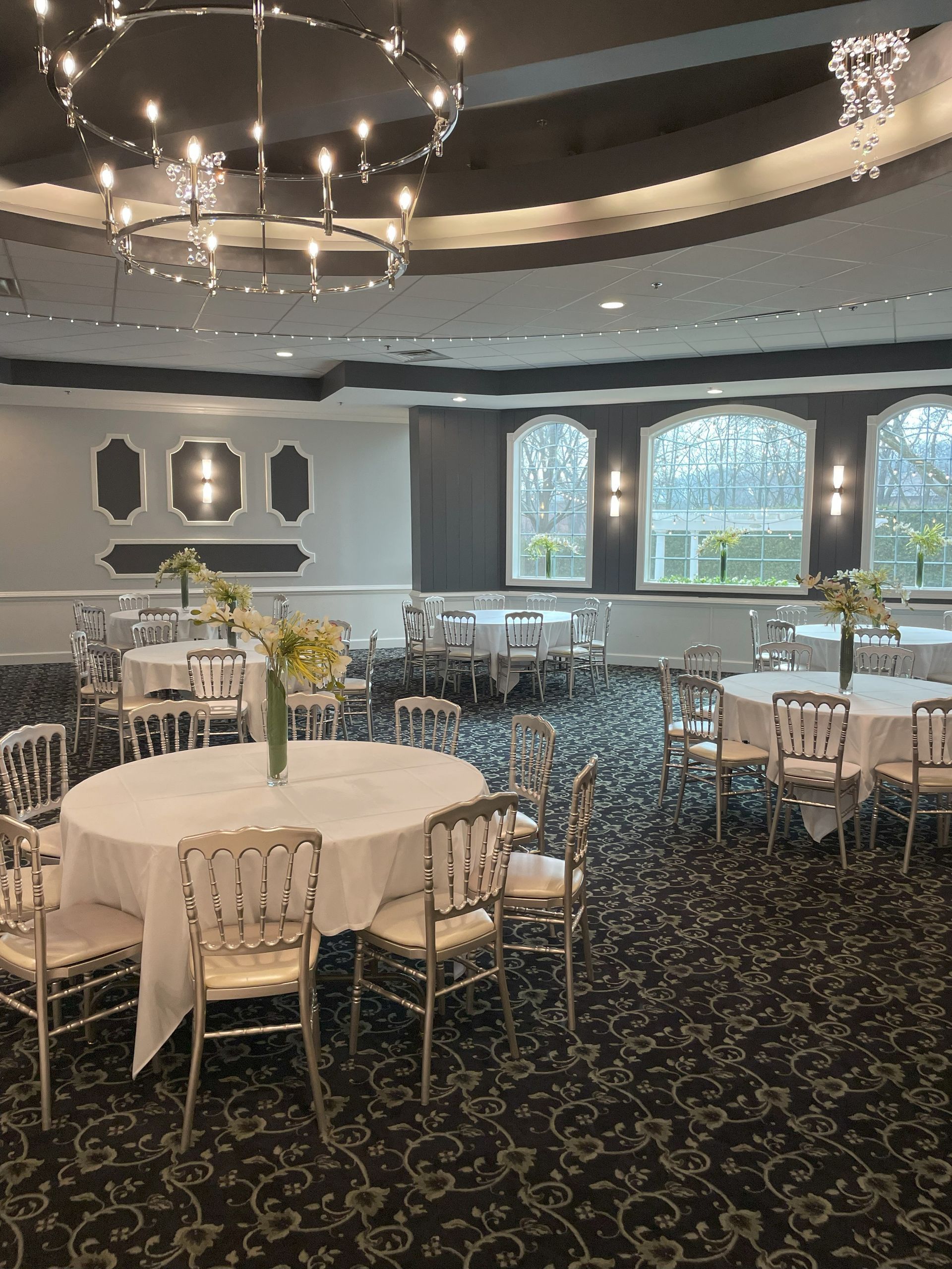 Elegant banquet hall with round tables, white chairs, and chandeliers by large arched windows