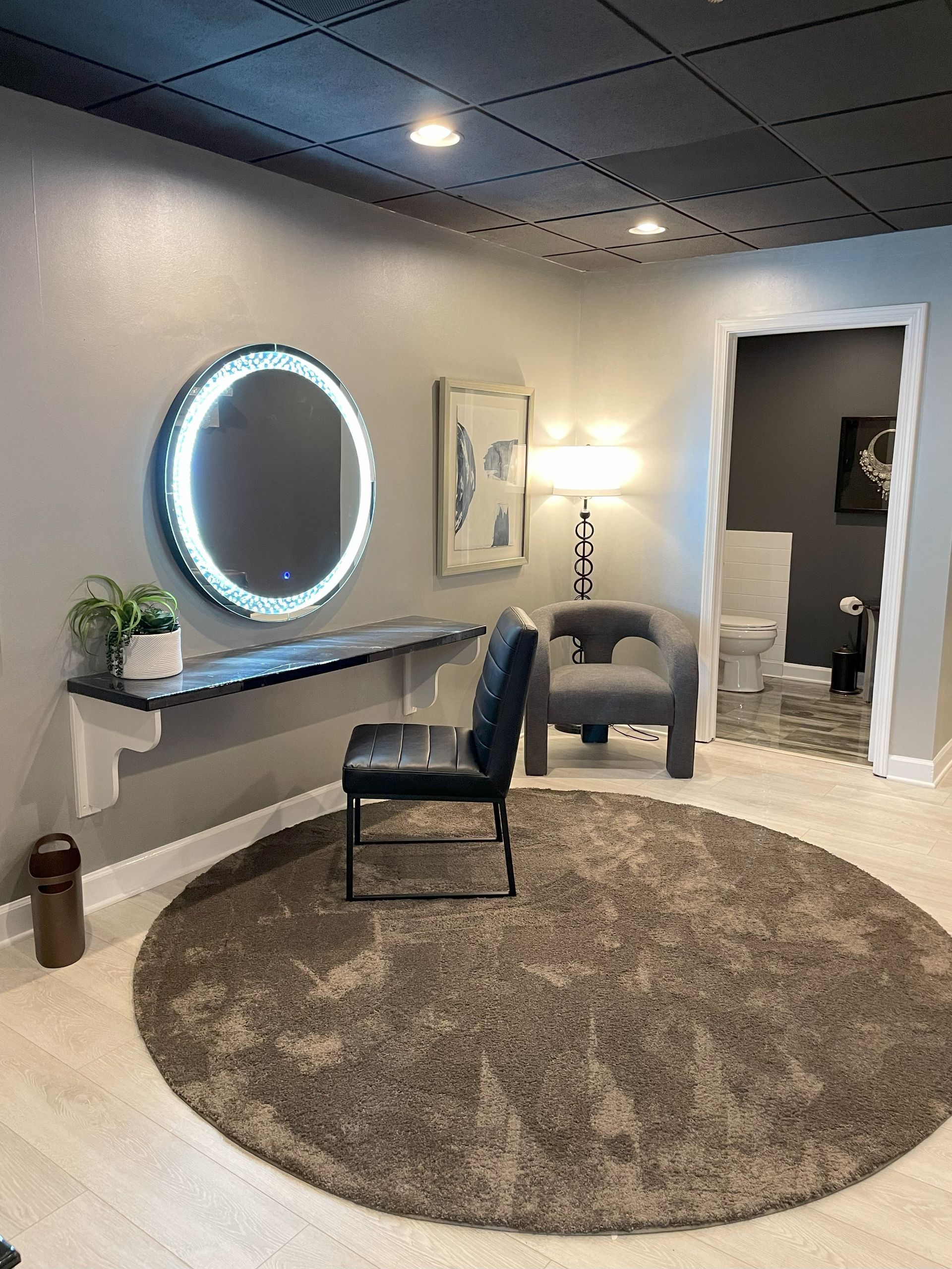 Modern waiting room with round lit mirror, chairs, small table, rug, and doorway to adjoining room