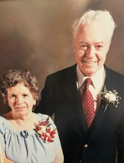 Smiling older couple in formal attire, posed against a plain studio backdrop.