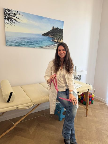 Una persona sorridente con indosso una giacca a fantasia è seduta su una poltrona color crema, appoggiata a una parete bianca e verde chiaro.