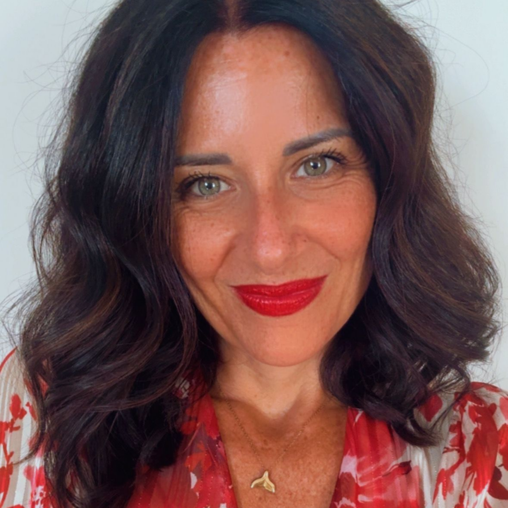 In una stanza con degli scaffali, una persona dai lunghi capelli scuri sorride indossando un blazer rosso acceso e un dolcevita nero.