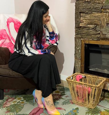 Una persona con lunghi capelli scuri è seduta su una sedia, indossa una blusa a fiori, una gonna nera e scarpe con tacco sfumate, accanto a un cesto di vimini.