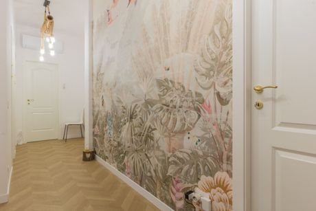 Un corridoio con pavimento a spina di pesce in legno chiaro, un murale raffigurante foglie tropicali, una lampada a sospensione e porte a pannelli bianchi.