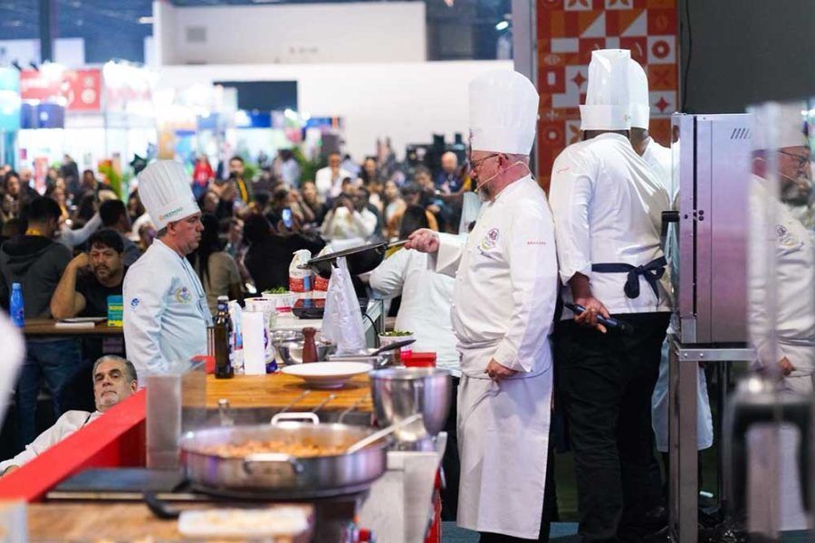 Chefs de uniforme branco e chapéus altos preparam comida em uma cozinha aberta durante um evento movimentado, com o público ao fundo.