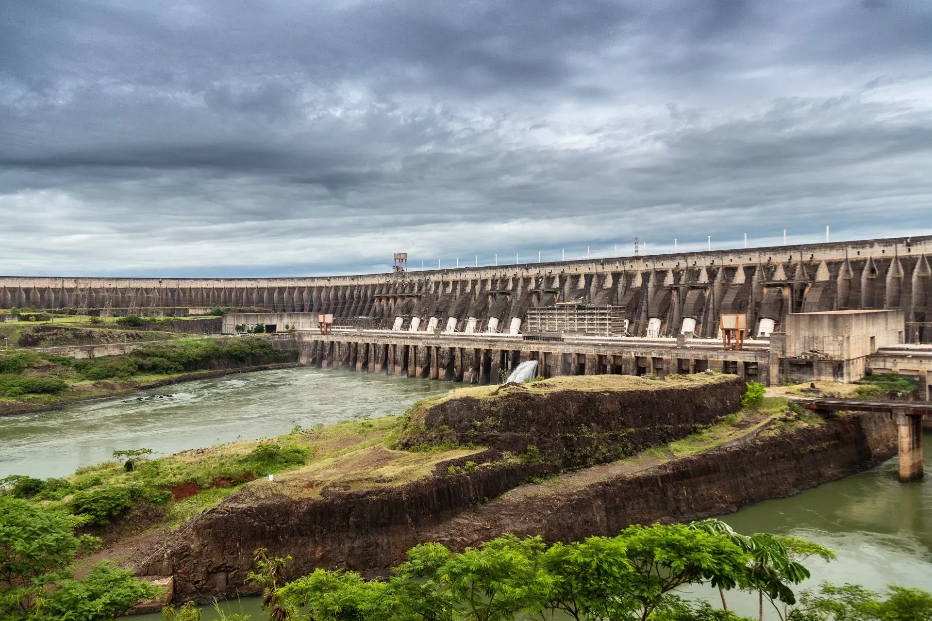 Uma grande represa com várias comportas de água sob um céu nublado, em uma paisagem verdejante.