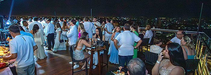 Uma festa animada em um terraço ao ar livre à noite, com pessoas em pé e sentadas em mesas com vista para o horizonte da cidade.