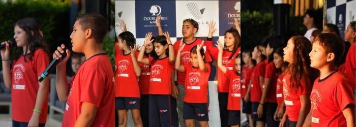Um grupo de pessoas vestindo camisetas vermelhas iguais canta em microfones em um palco em frente a um painel de fundo com a marca da empresa.