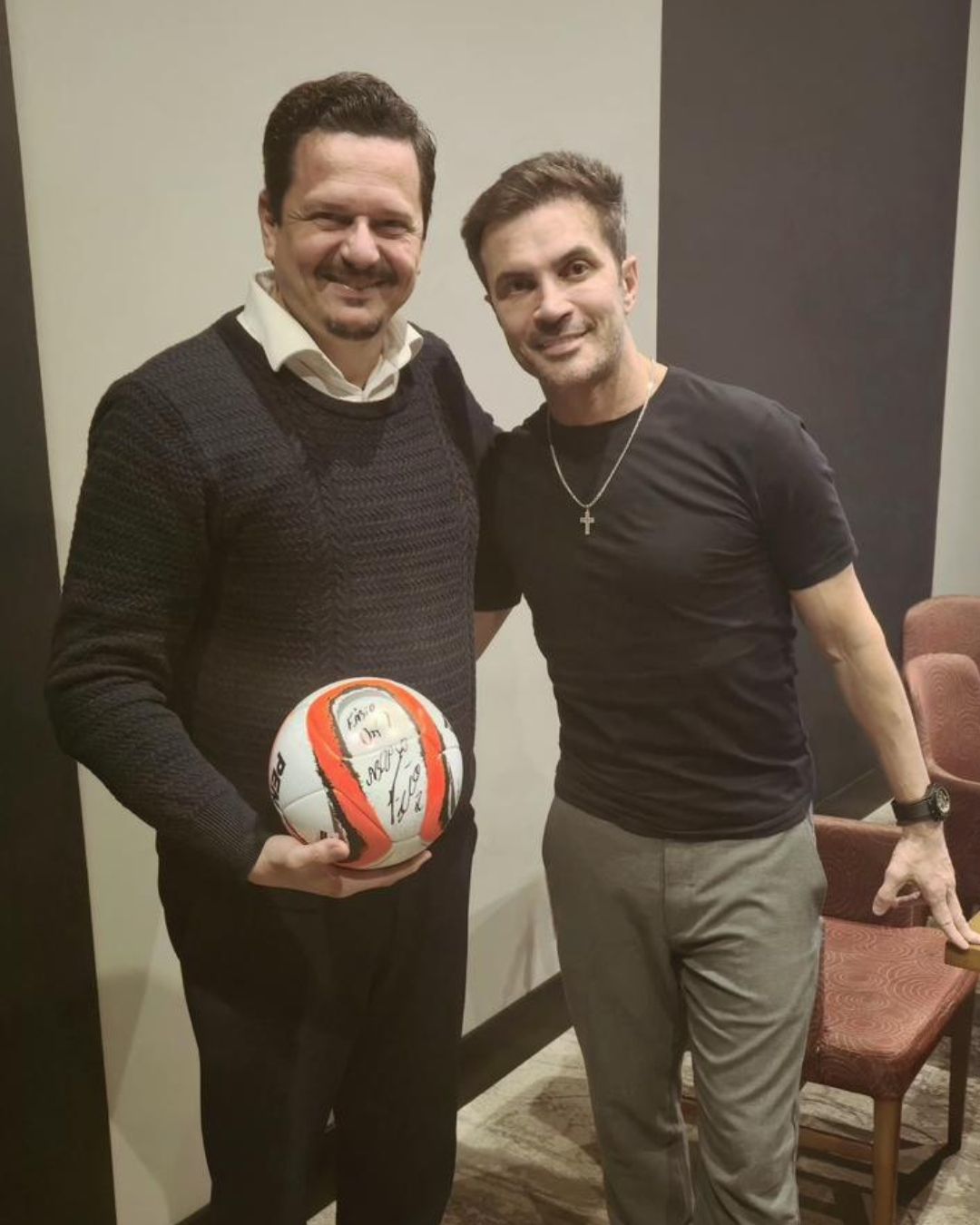 Dois homens estão lado a lado; um deles segura uma bola de futebol autografada, ambos sorrindo em um ambiente interno com cadeiras.