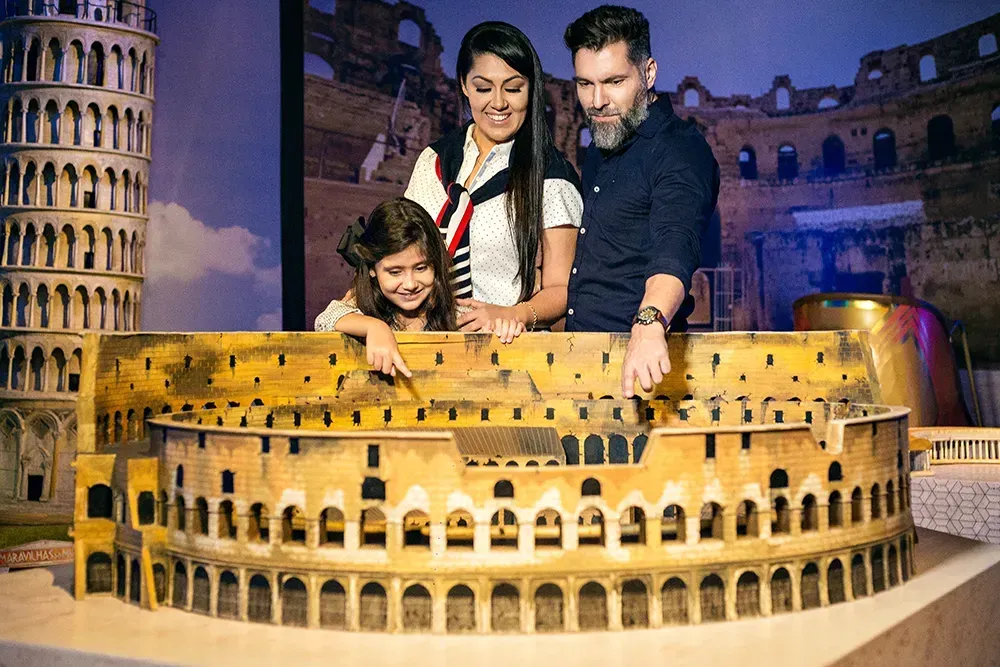 Família aponta para um Coliseu em miniatura, com a Torre Inclinada de Pisa ao fundo.