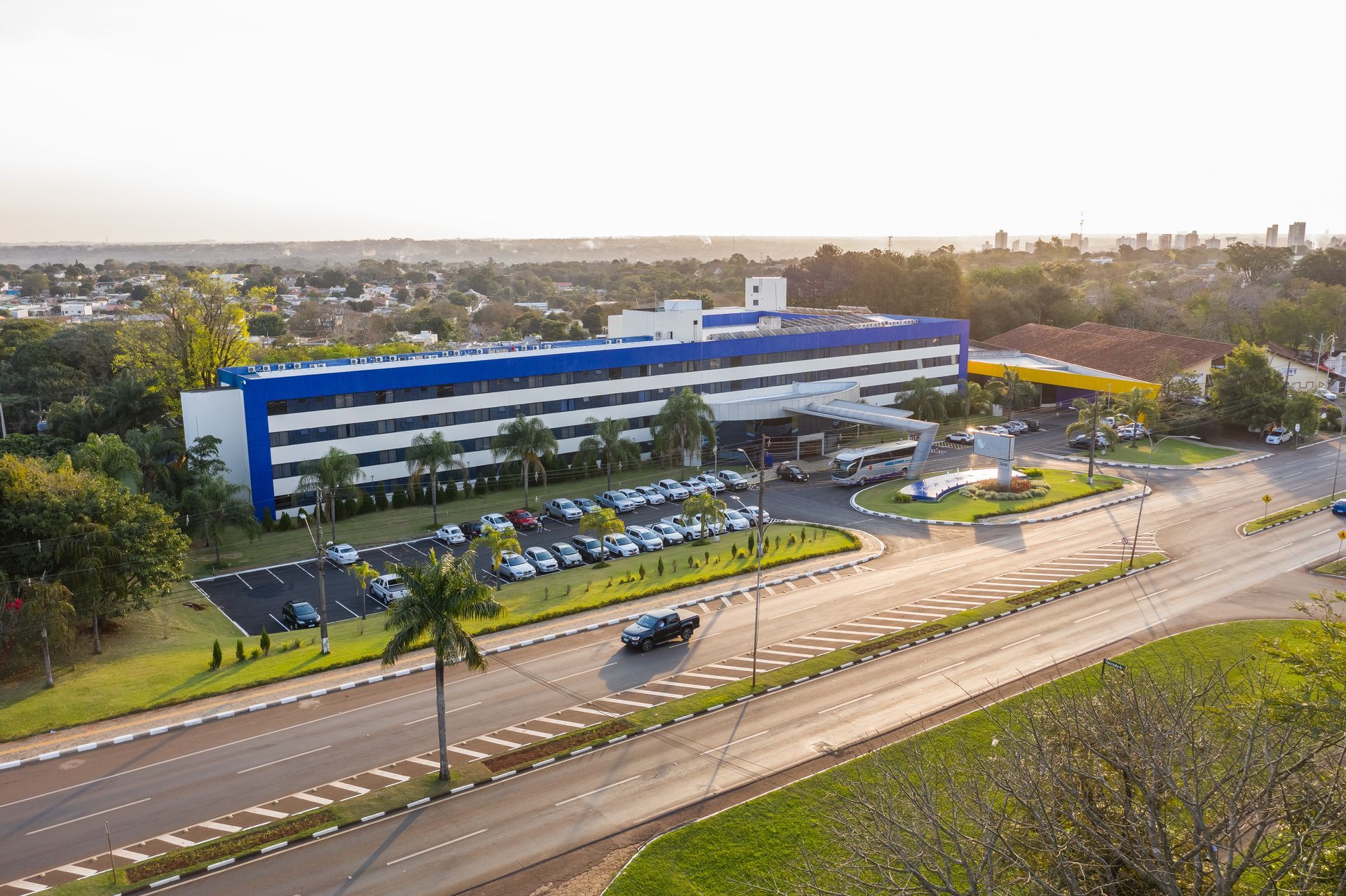 Vista aérea de um hotel de vários andares com detalhes externos em azul e branco, cercado por estacionamento e vegetação próximo a uma estrada.