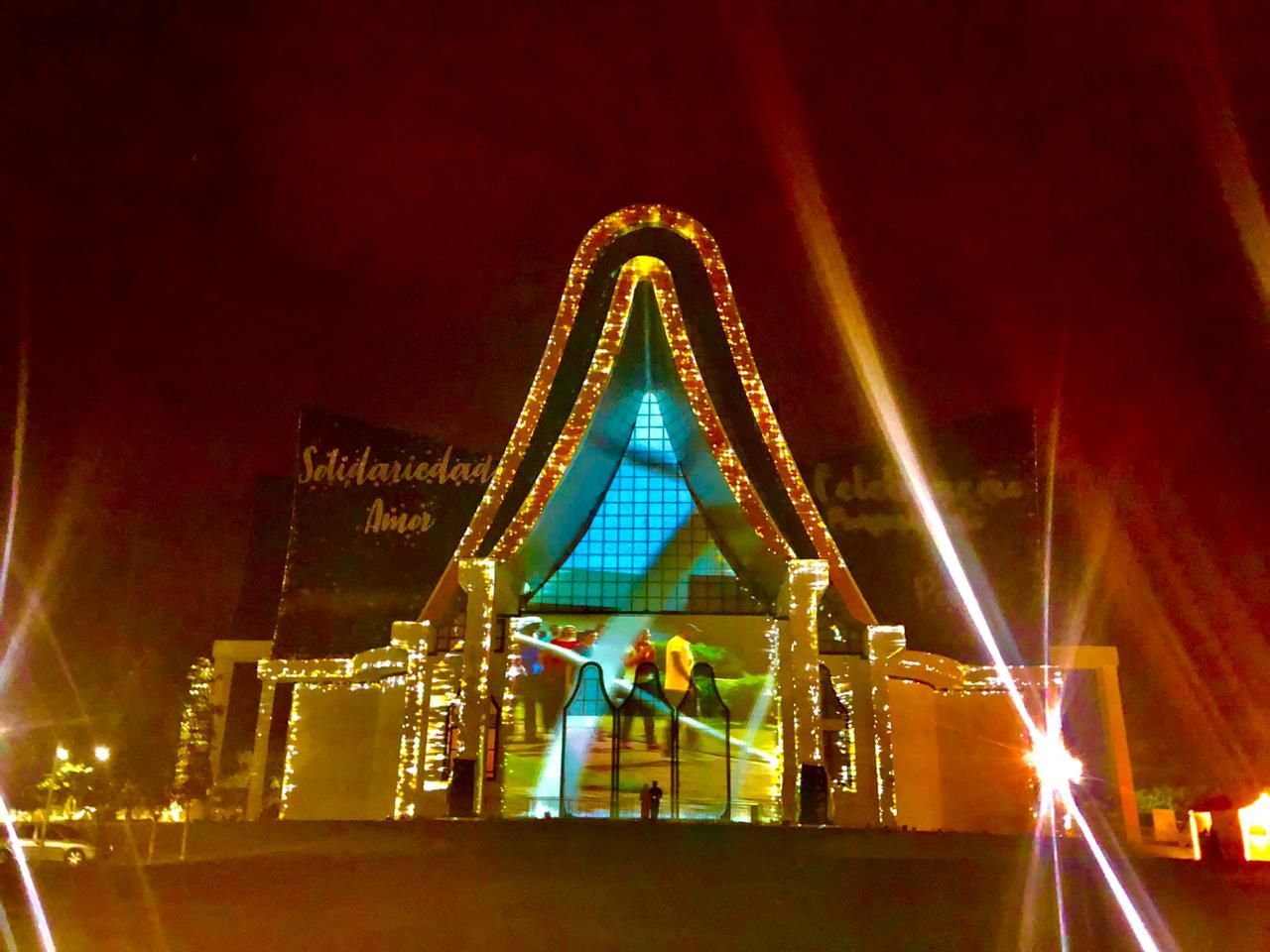 Igreja à noite, decorada com luzes e projeção mapeada. O texto 