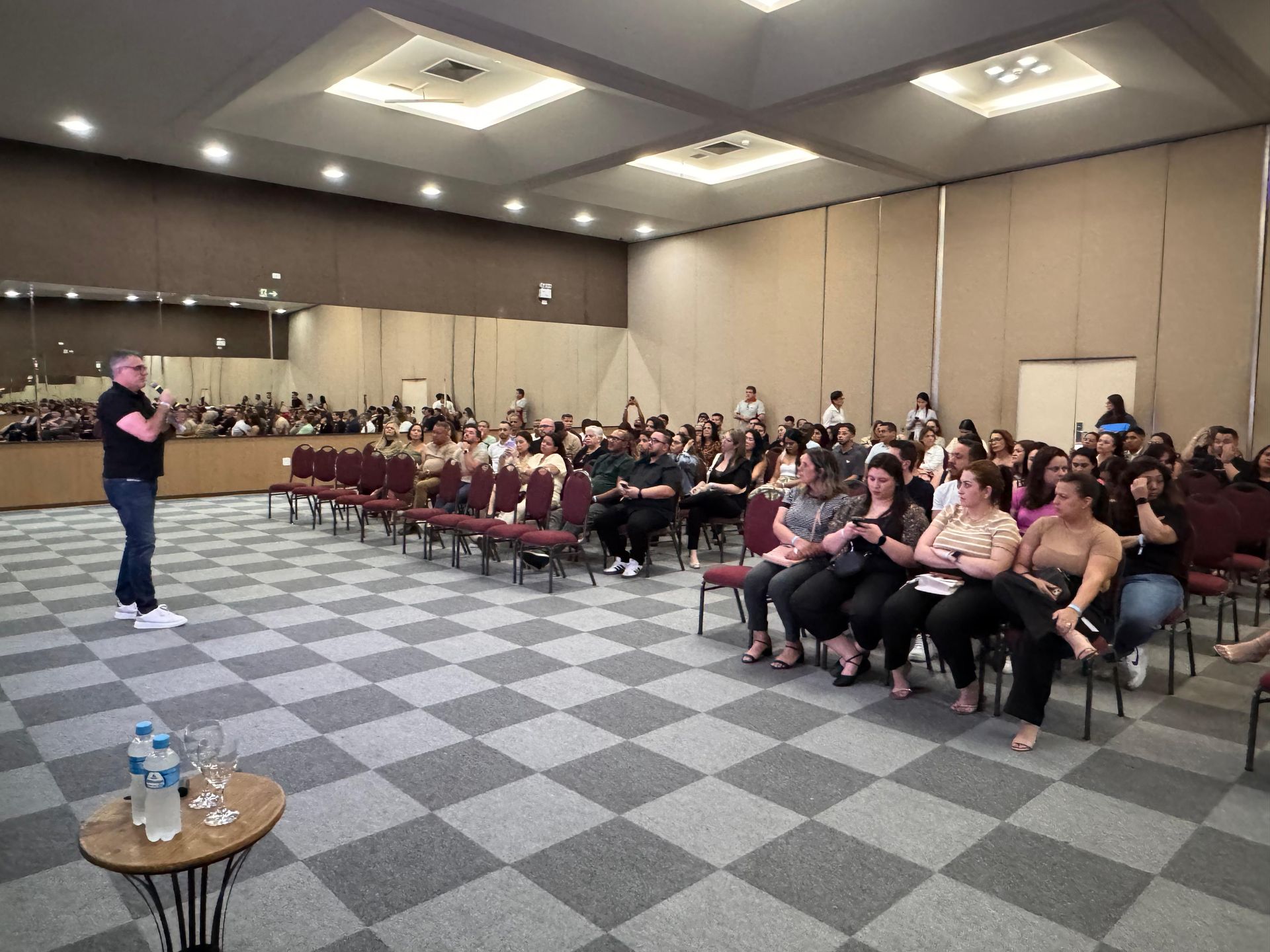 Uma pessoa está de pé, apresentando para uma plateia sentada em cadeiras em uma grande sala de conferências com carpete.