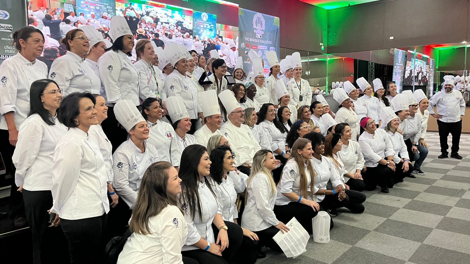 Um grande grupo de chefs com uniformes brancos posa junto em um palco para uma foto de grupo durante um evento.