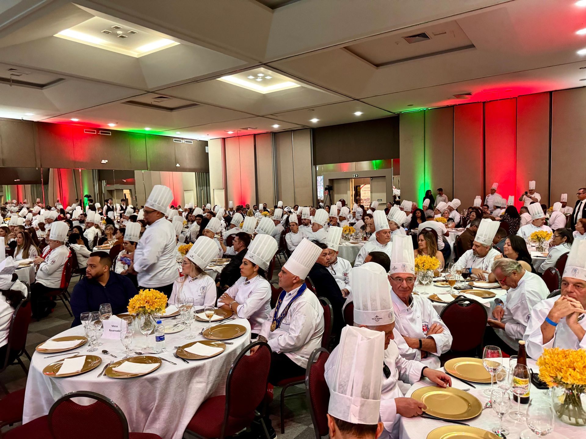 Vários chefs de uniforme branco e chapéu alto estavam sentados às mesas em um grande salão de eventos com iluminação de destaque em vermelho e verde.