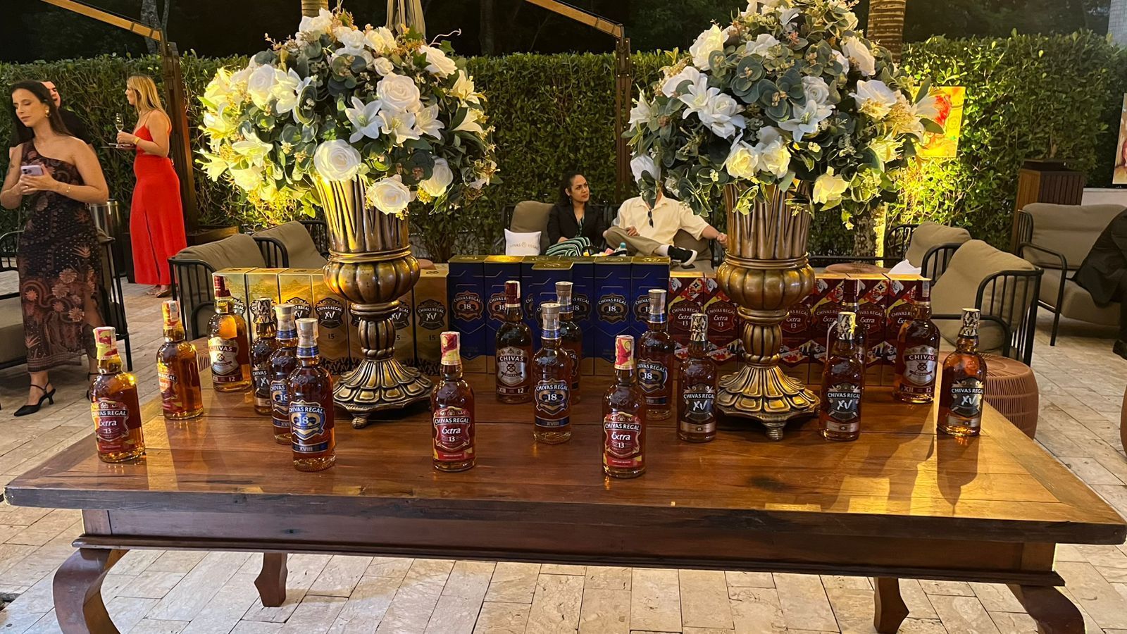 Uma mesa de madeira exibe arranjos florais decorativos e garrafas de bebidas alcoólicas em um evento noturno ao ar livre.