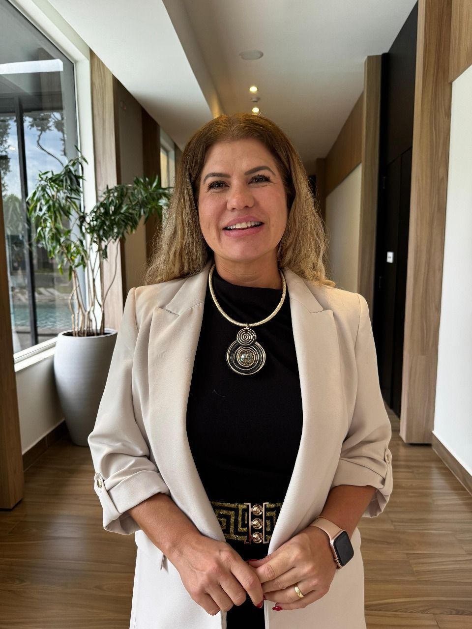 Uma pessoa sorridente, vestindo um blazer creme e uma blusa preta, com um colar chamativo grande, está em um corredor bem iluminado.
