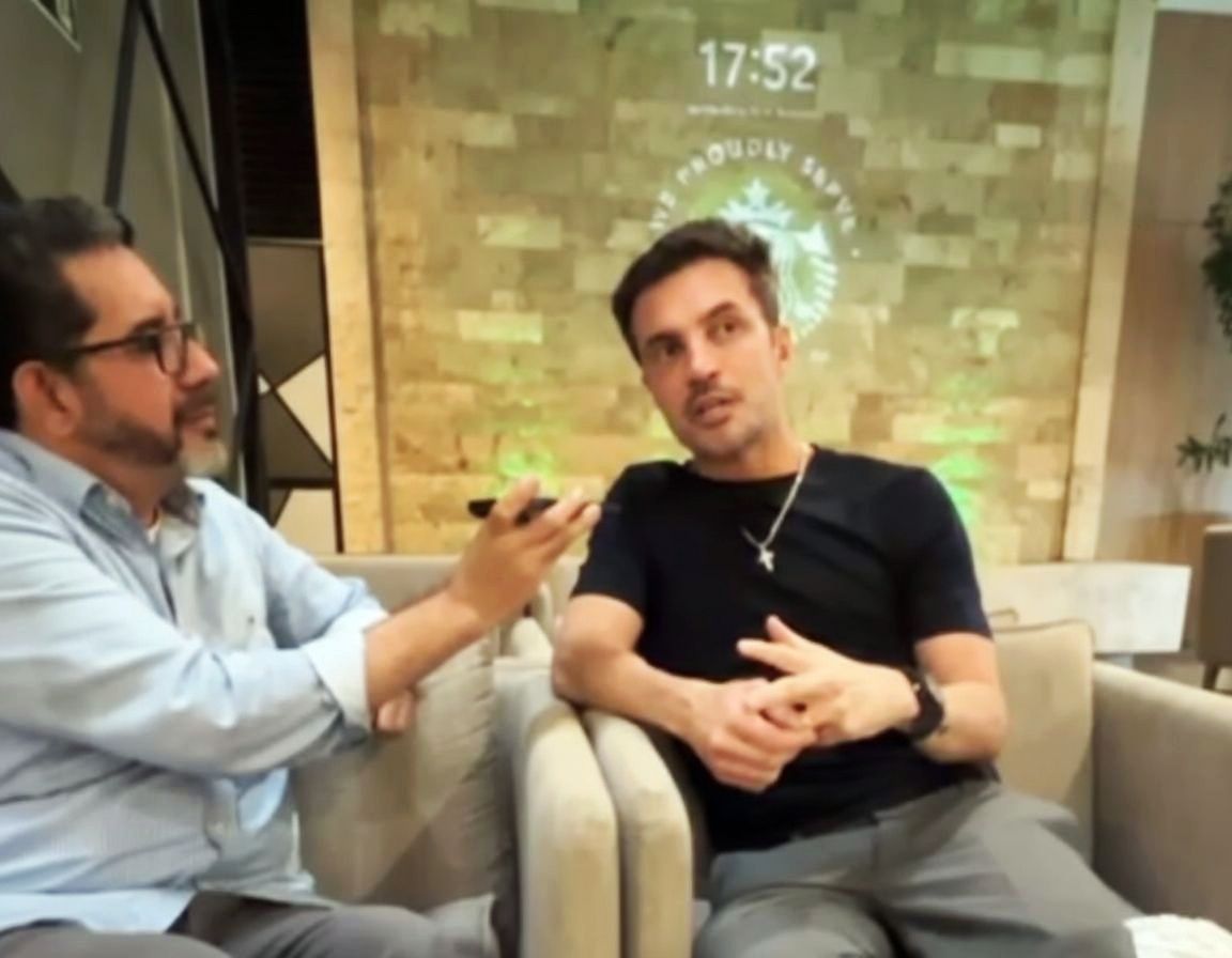 Um entrevistador segura um microfone em direção a uma pessoa que está falando, enquanto esta está sentada em uma área de estar moderna.