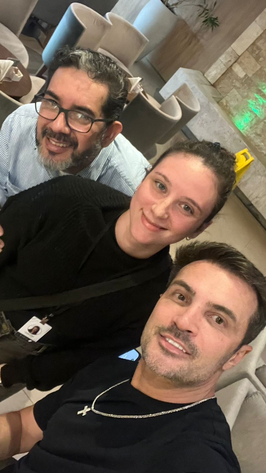 Uma selfie de três pessoas sorridentes, duas com camisas pretas e uma com uma camisa azul-clara estampada, em uma área de descanso.