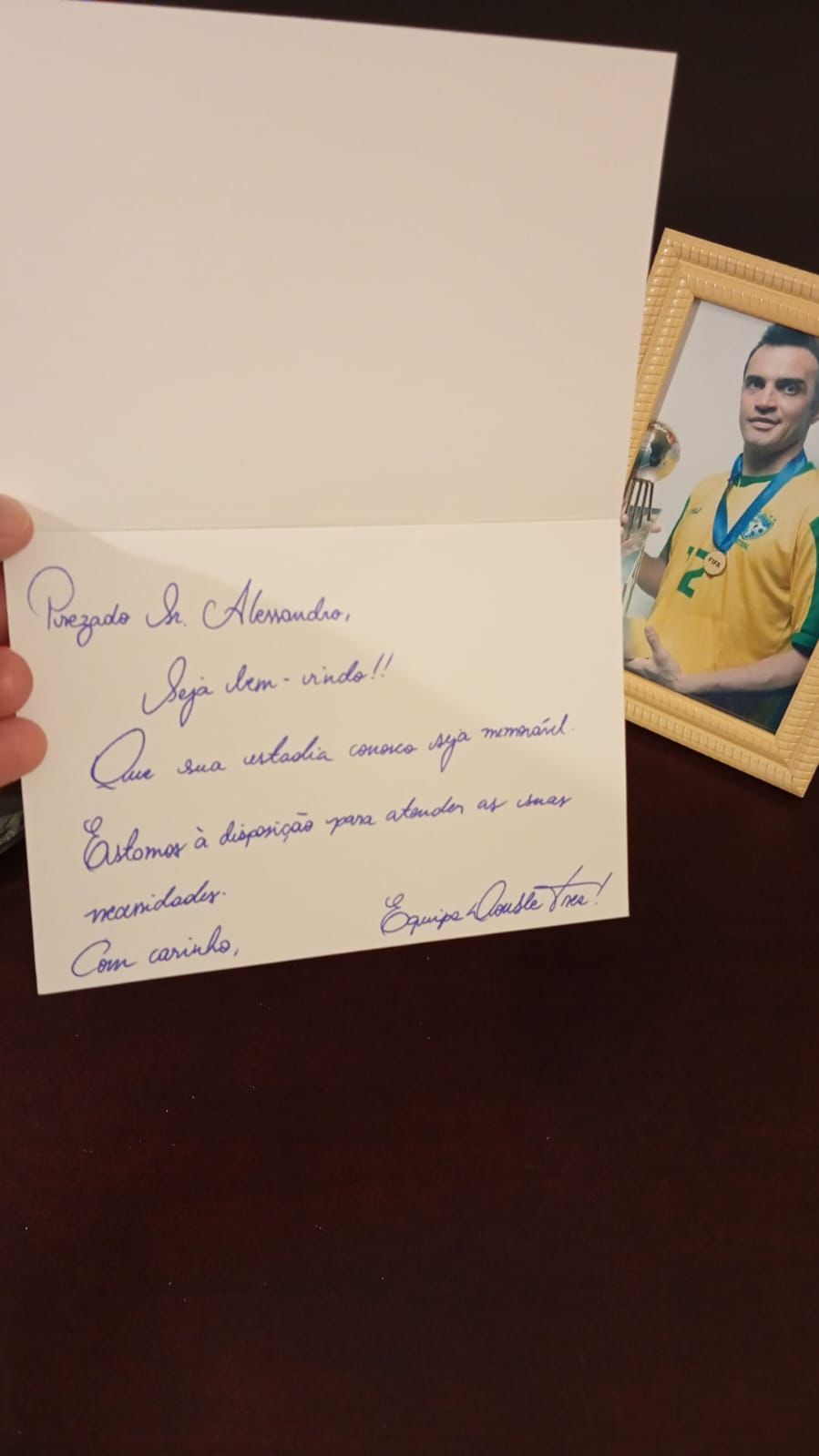 Uma mão segura um bilhete de boas-vindas escrito à mão ao lado de uma foto emoldurada de uma pessoa vestindo uma camisa amarela e uma medalha.