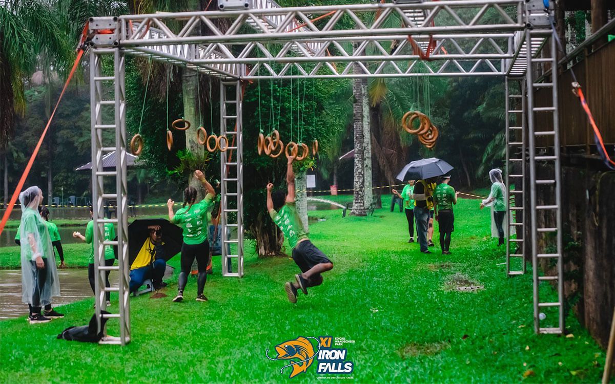 Os participantes, vestindo camisetas verdes, percorrem uma pista de obstáculos ao ar livre, com uma pessoa suspensa por argolas sob uma estrutura metálica.