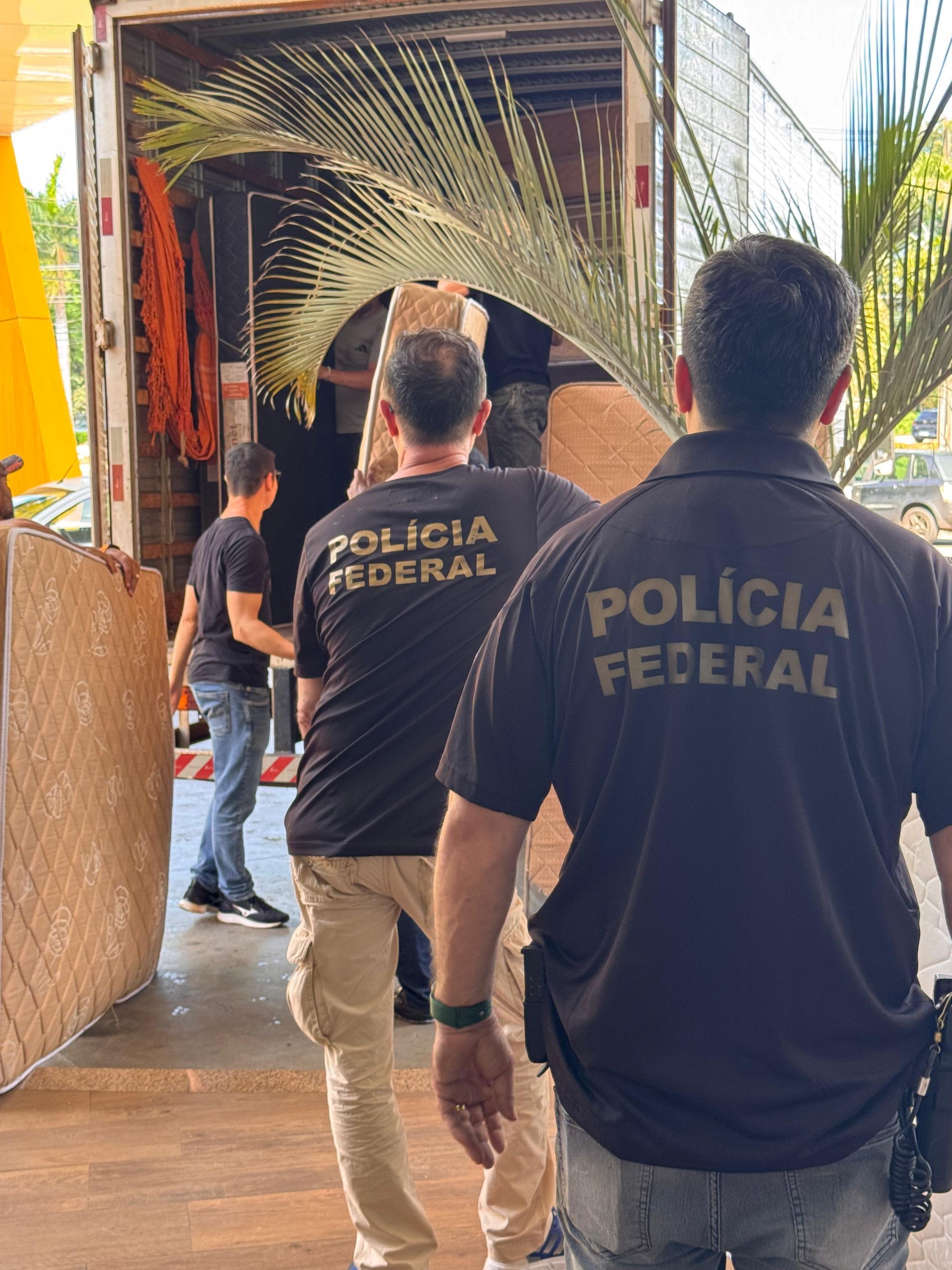 Dois agentes da Polícia Federal, vestindo camisas pretas com a marca da corporação, supervisionam o carregamento de um caminhão contendo folhagem e carga.