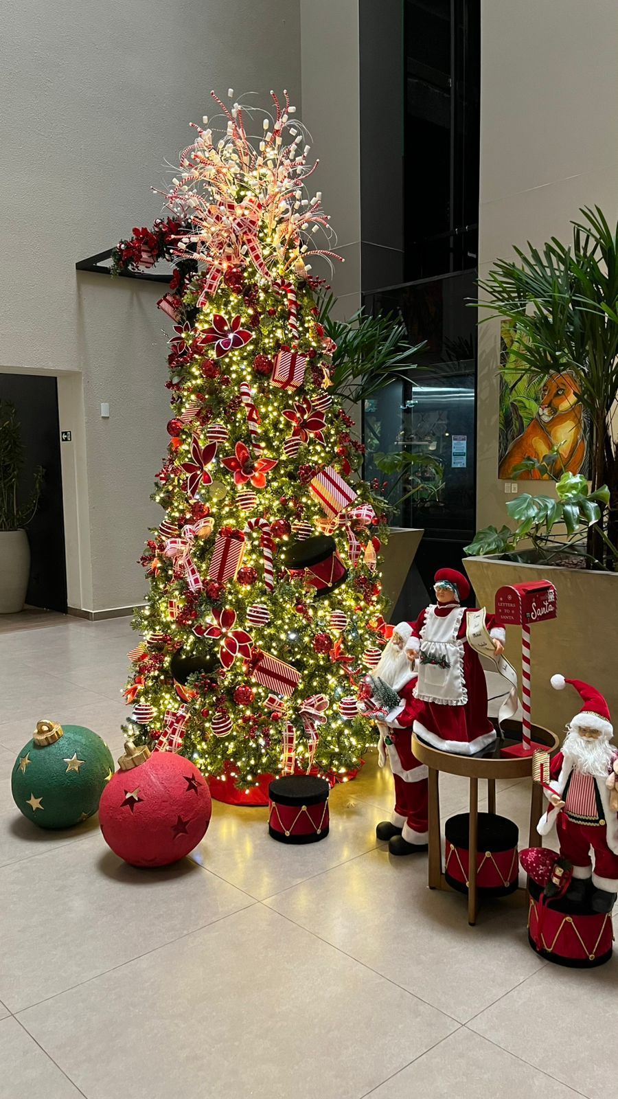 Uma árvore de Natal alta e iluminada, adornada com enfeites vermelhos e dourados, fica ao lado de figuras decorativas do Papai Noel em um saguão.