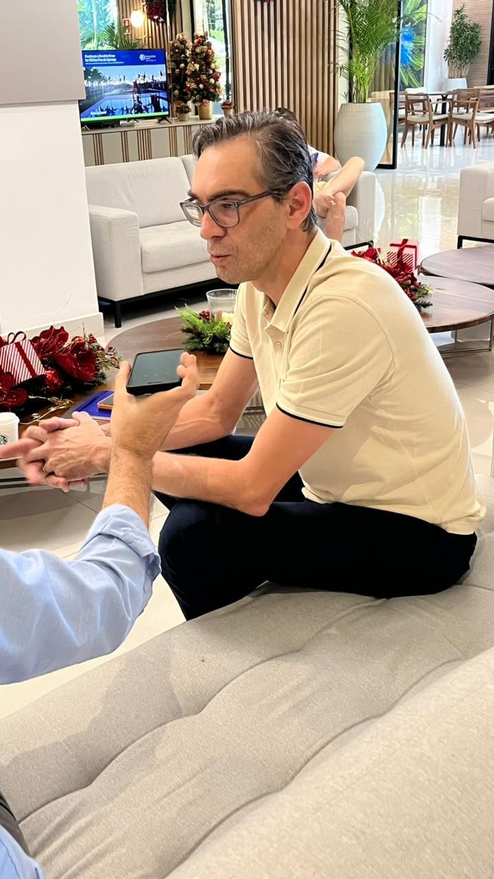 Um homem vestindo uma camisa polo creme está sentado em um sofá, conversando com alguém fora do enquadramento da câmera, que segura um smartphone para gravar a entrevista.