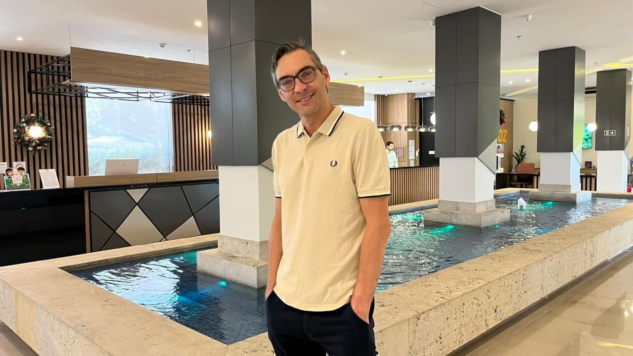 Uma pessoa vestindo uma camisa polo creme está sorrindo no saguão de um hotel, ao lado de uma fonte de pedra interna com colunas de sustentação escuras.
