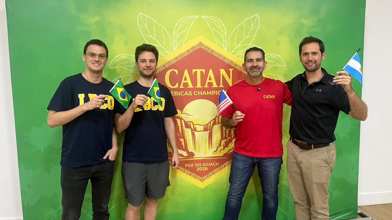 Quatro pessoas estão em pé diante de uma faixa de campeão do CATAN, cada uma segurando uma pequena bandeira nacional.