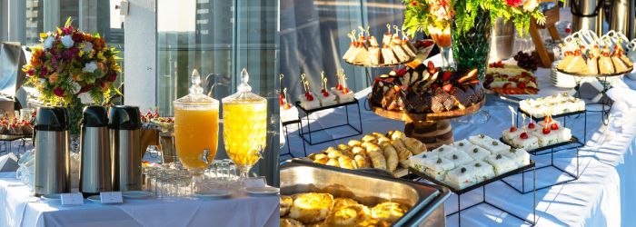 Uma mesa de bufê com jarras de suco, diversas travessas de doces, aperitivos e um arranjo floral como centro de mesa.