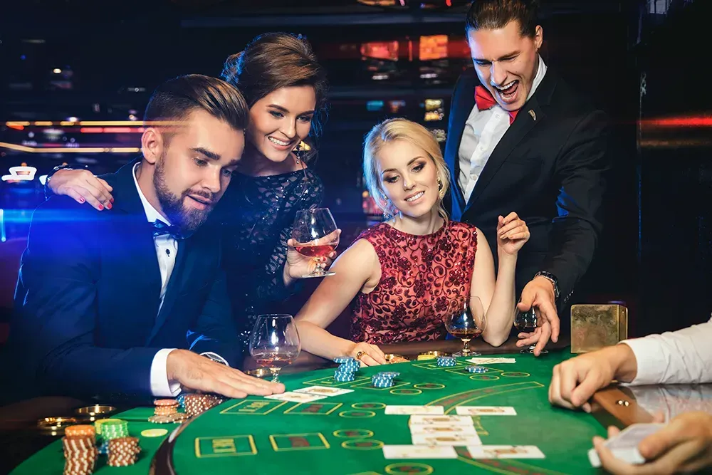 Grupo de amigos em uma mesa de cassino, jogando cartas. Expressões alegres, traje formal, feltro verde, fichas e bebidas.