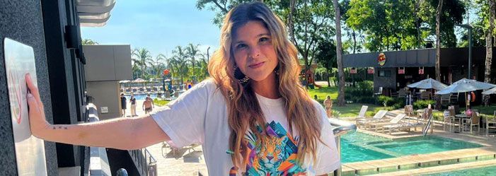 Uma pessoa jovem de cabelos ondulados encosta-se numa parede, vestindo uma camiseta branca casual, com vista para a área ensolarada da piscina de um resort.