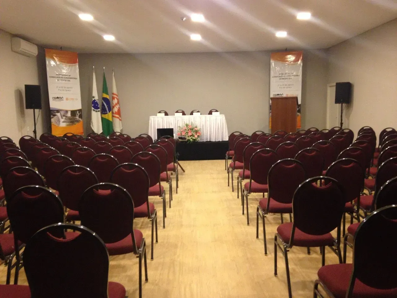 Fileiras de cadeiras vermelhas ficam de frente para um palco com uma mesa e um pódio em uma sala de conferências.